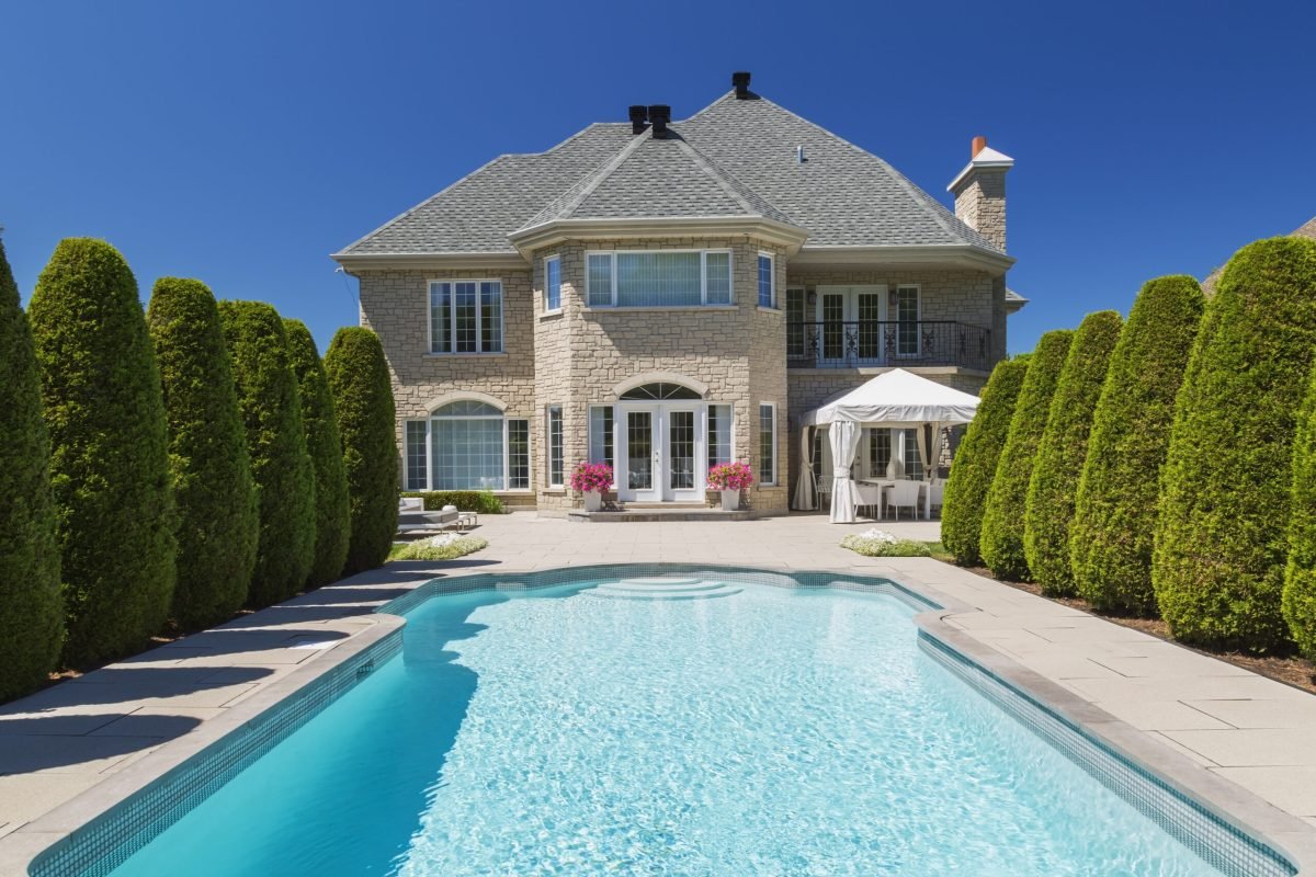 Outdoor swimming pool bordered by rows of cedar trees (Thuja occidentallis) at rear of luxurious house with beige stone